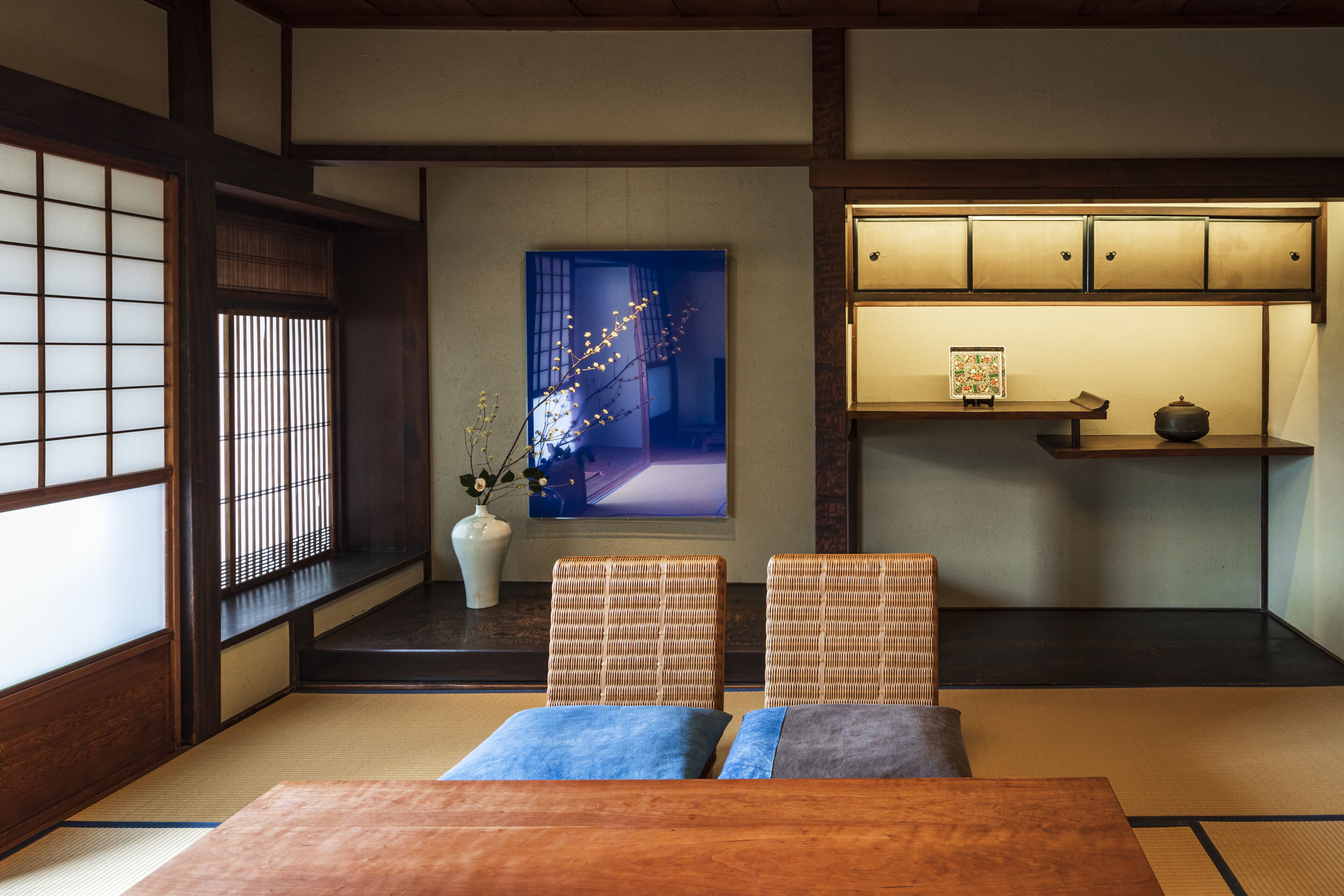 Una tradizionale stanza giapponese con pavimento in tatami, un tavolino basso, due angoli salotto imbottiti, un vaso decorativo e una mensola a parete con una cornice e una piccola pianta.