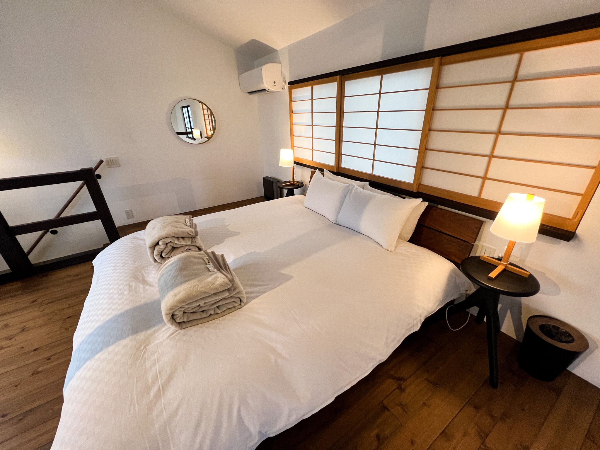 Une chambre d’hôtel avec un grand lit, une table de chevet avec une lampe, un miroir rond et une tête de lit en bois avec une fenêtre.