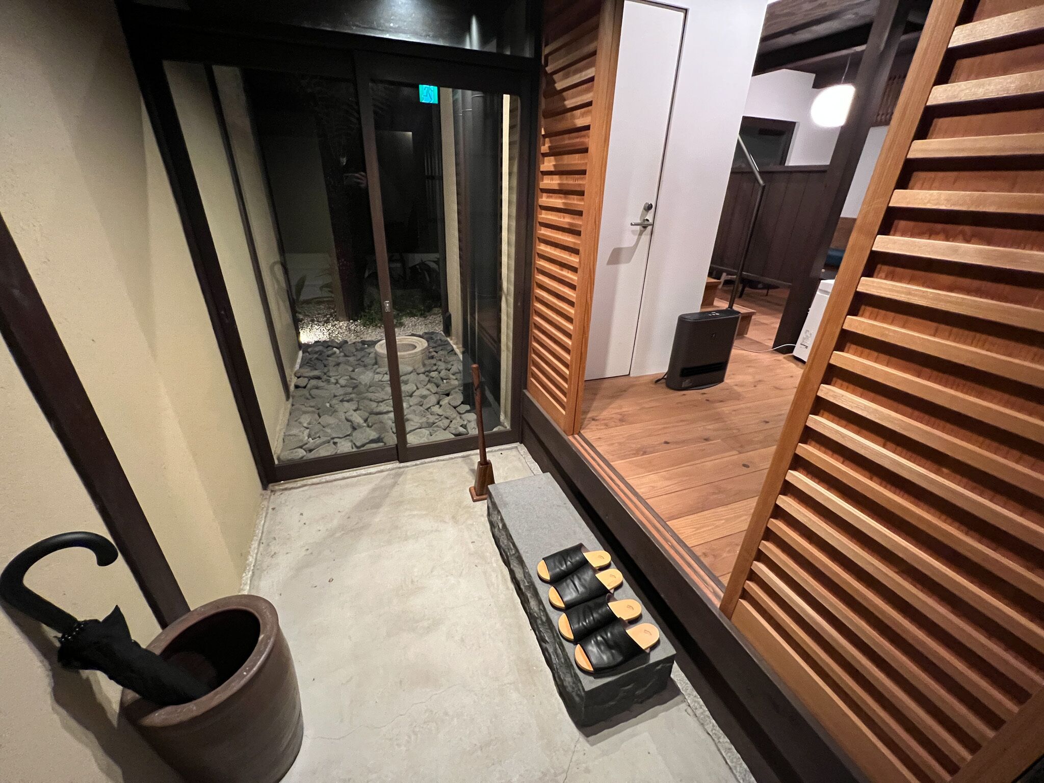 A modern interior with a sliding glass door leading to a stone-tiled area, a wooden bench with shoes, and a black trash bin.