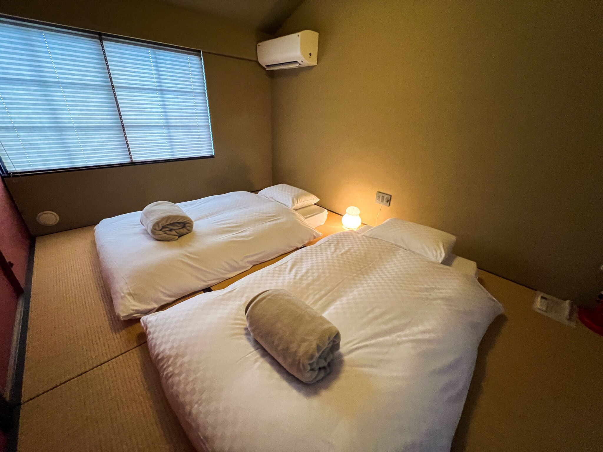 A small, single-room hotel room with two beds, a window with blinds, a wall-mounted air conditioner, and a bedside lamp.