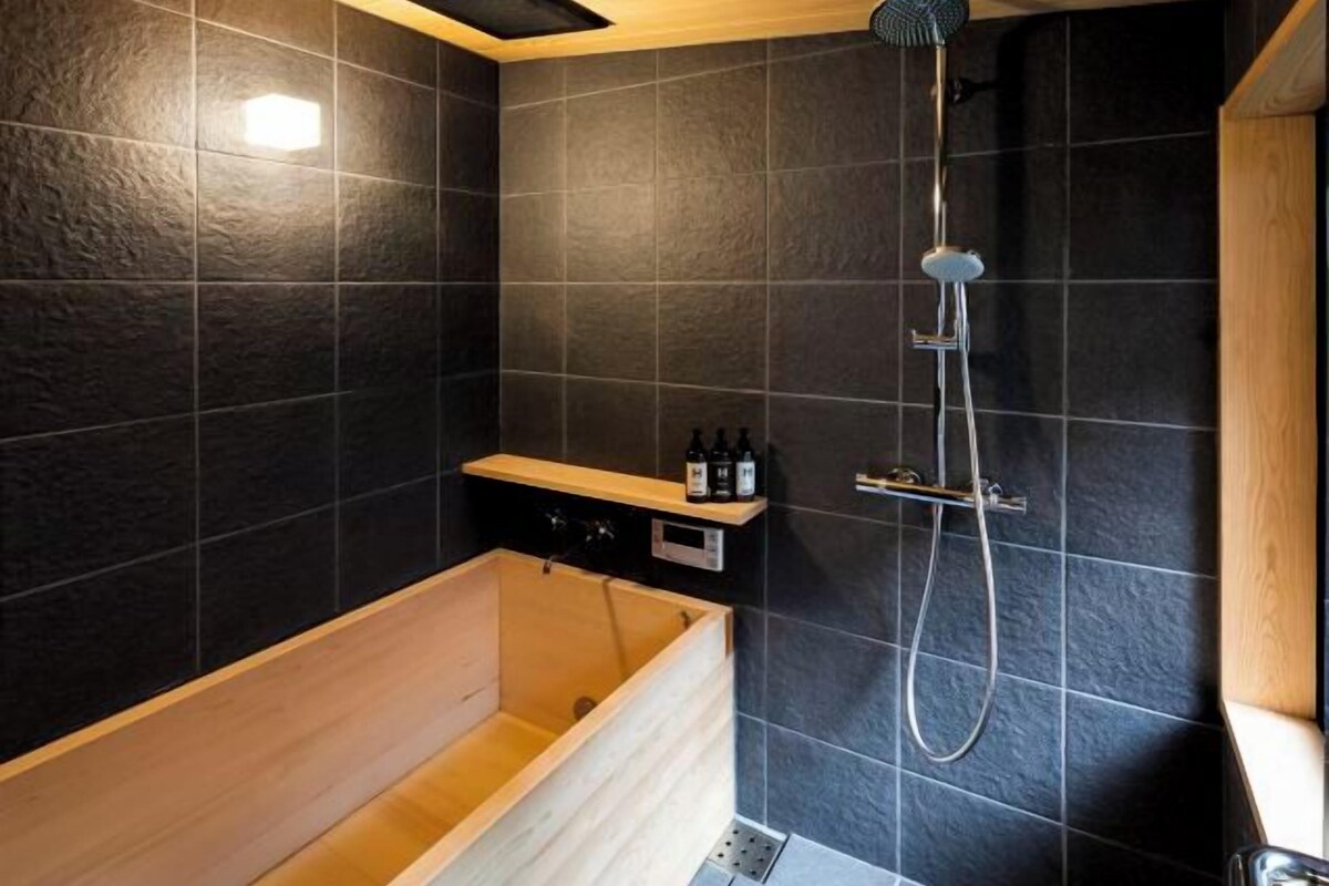 A modern bathroom with a wooden bathtub, a shower with a handheld showerhead, and a shelf with toiletries.