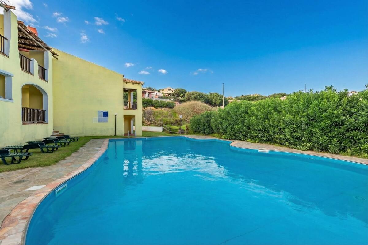 Une piscine bordée de pierres, un bâtiment doté d’un balcon et une haie en arrière-plan.