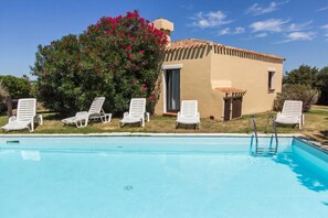 Uma piscina com cadeiras de lounge brancas, um pequeno edifício com telhado em azulejo e uma árvore com flores vermelhas.