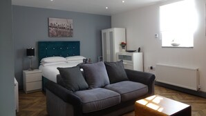 Een moderne slaapkamer met een bed, een grijze bank, een houten salontafel, een witte garderobe, een wit kastje en een raam met natuurlijk licht.