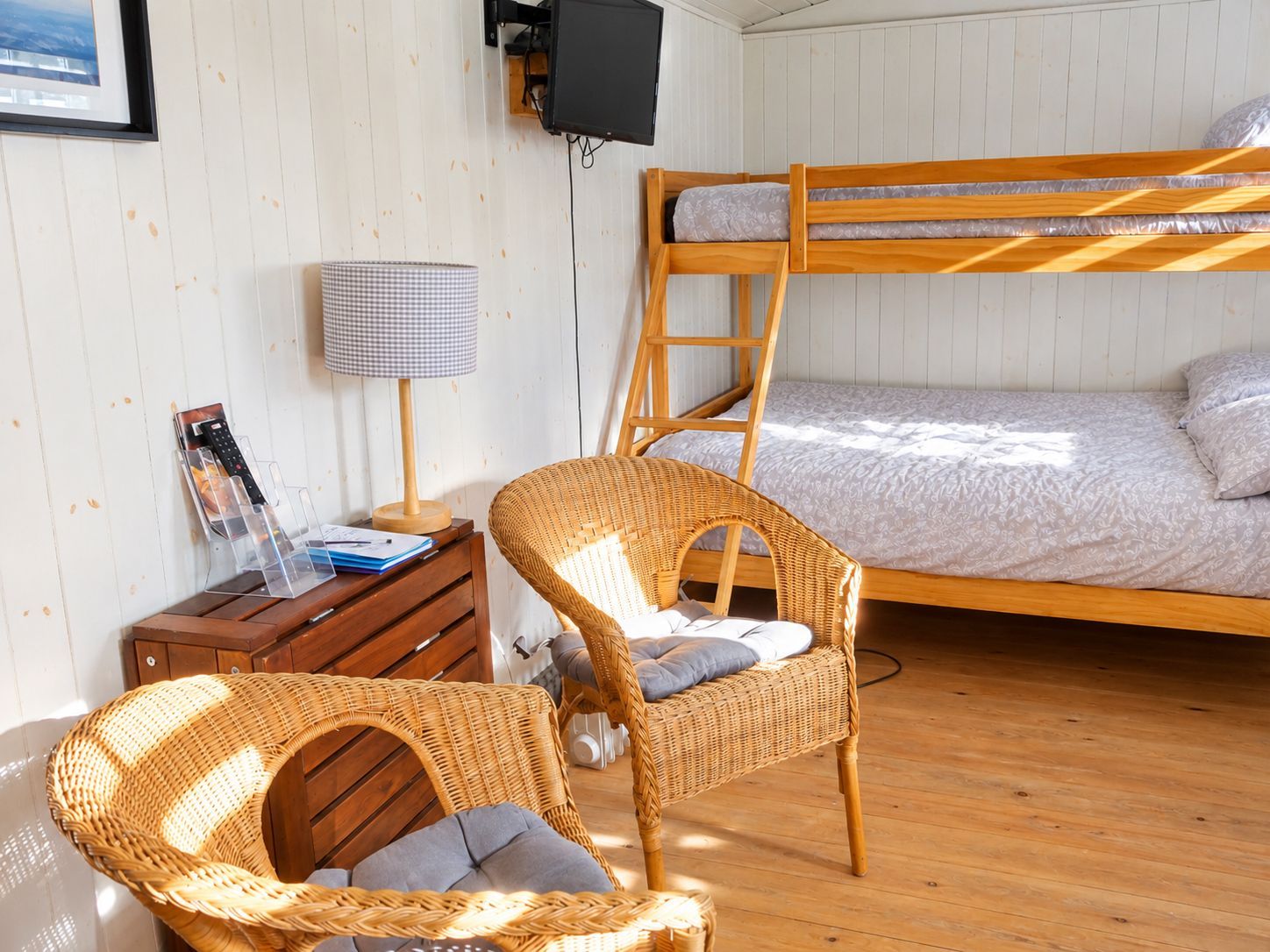 A bunk bed with a wooden frame, a wicker chair with cushions, a desk with a lamp, and a framed picture on the wall.