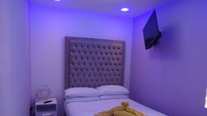 Une chambre à coucher avec une tête de lit capitonnée, un lit recouvert de draps blancs, une table de chevet avec une lampe et un téléviseur fixé au mur.