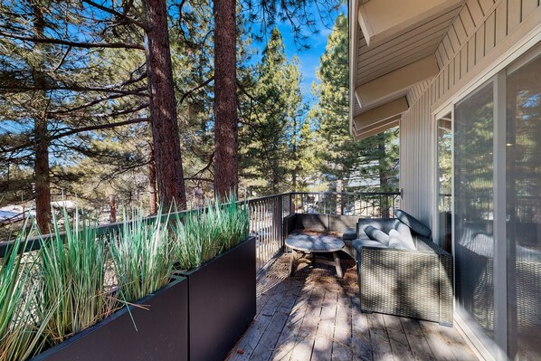 Relax on the outdoor deck with a wicker sofa and small table, surrounded by lush greenery and a black railing.