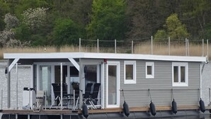 Ein schwimmendes Hausboot mit überdachter Terrasse, Fenstern und Geländer.