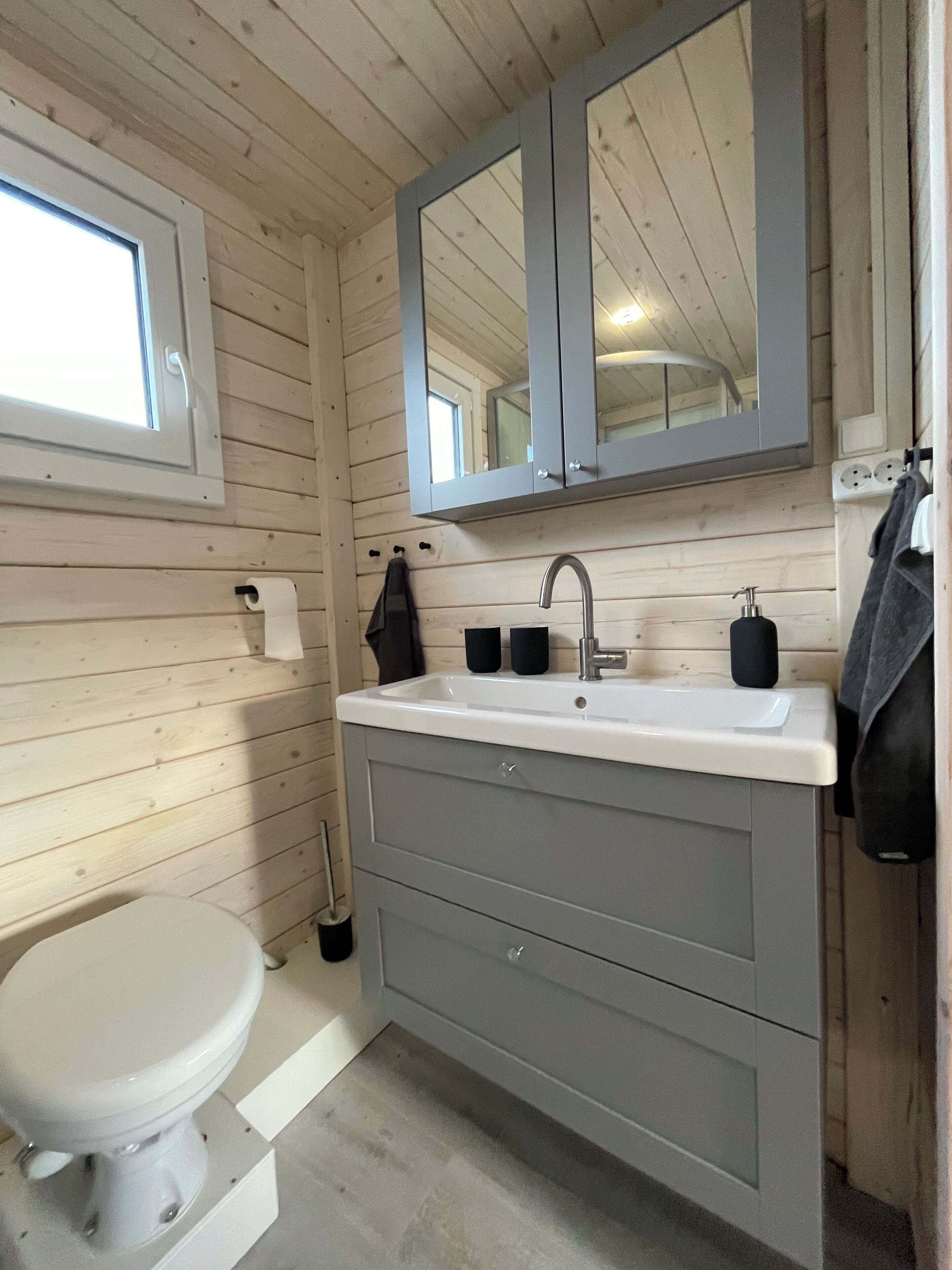 Ein kleines, kompaktes Badezimmer mit Holzwänden, einem weißen Waschbecken, einem Spiegel darüber und einer Toilette.