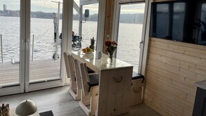 Eine Hütte mit Holzvertäfelung, einer Glastür, die zu einem Steg führt, einem Esstisch mit Stühlen und einem an der Wand befestigten Fernseher.