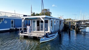 Ein Hausboot liegt in einer Marina festgemacht, mit einer überdachten Deckterrasse, einem kleinen Boot und einer Brücke im Hintergrund.