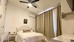 Un dormitorio con cama, mesita de noche, ventilador de techo, equipo de aire acondicionado, ventana con cortinas y baño con lavabo y espejo.