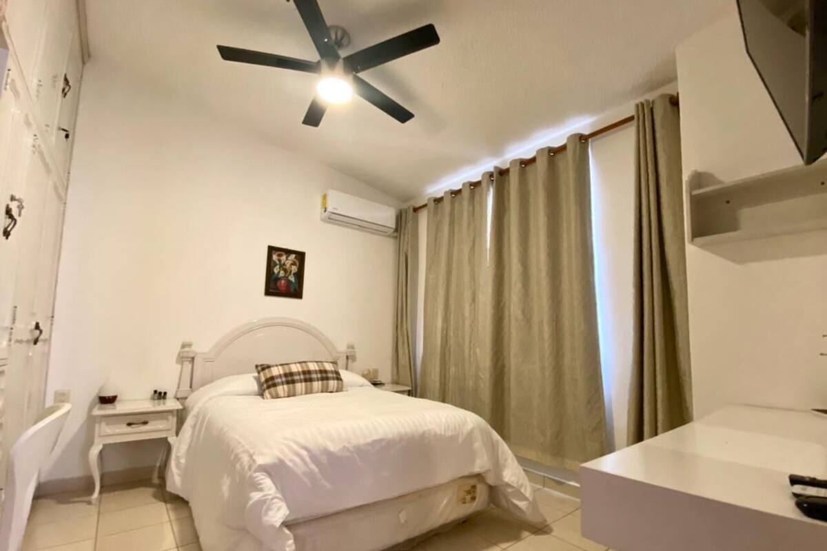 Un dormitorio con cama, mesita de noche, ventilador de techo, equipo de aire acondicionado, ventana con cortinas y baño con lavabo y espejo.
