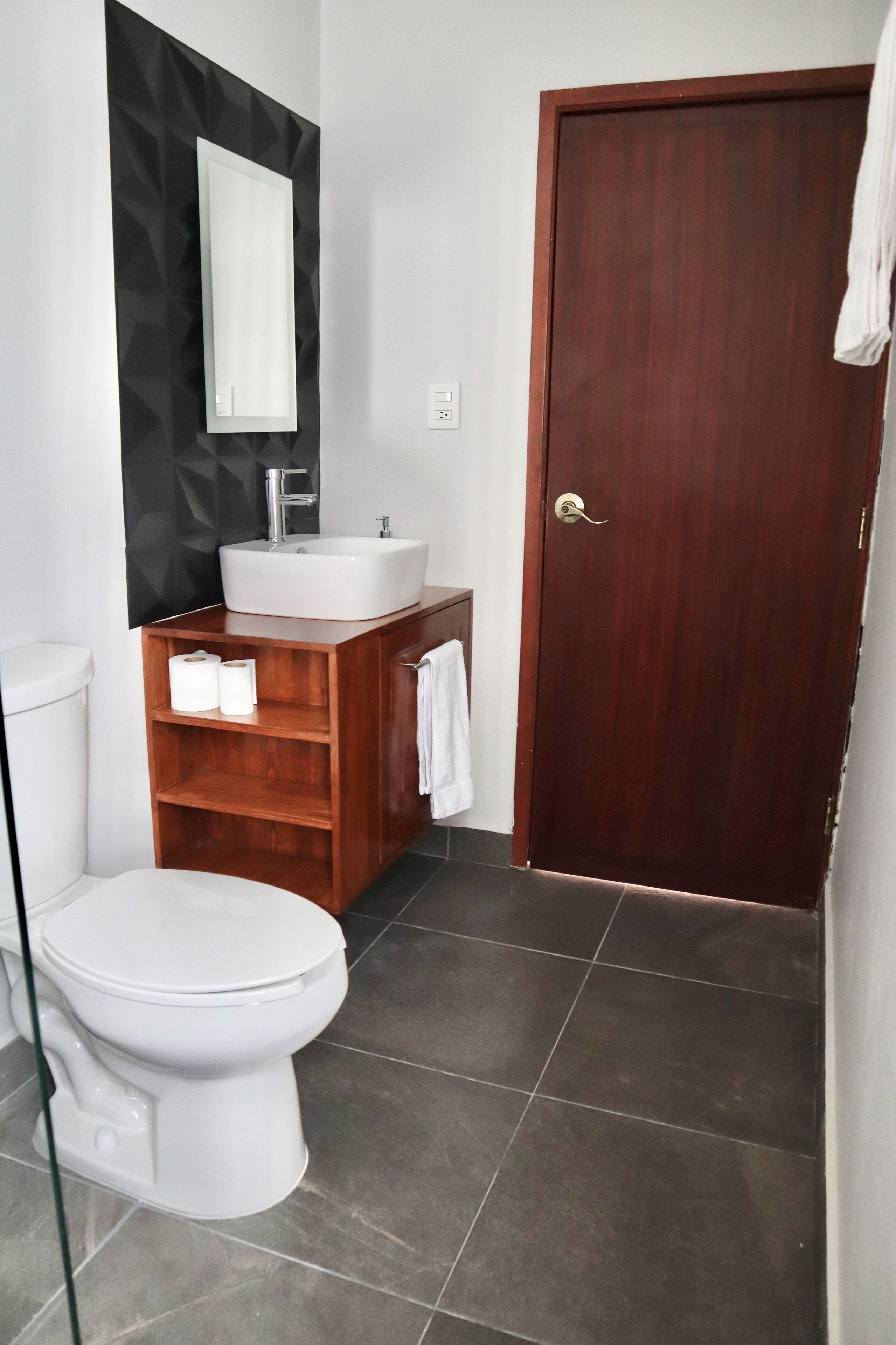 A modern bathroom with a white toilet, a wooden vanity with a sink, and a dark wooden door.