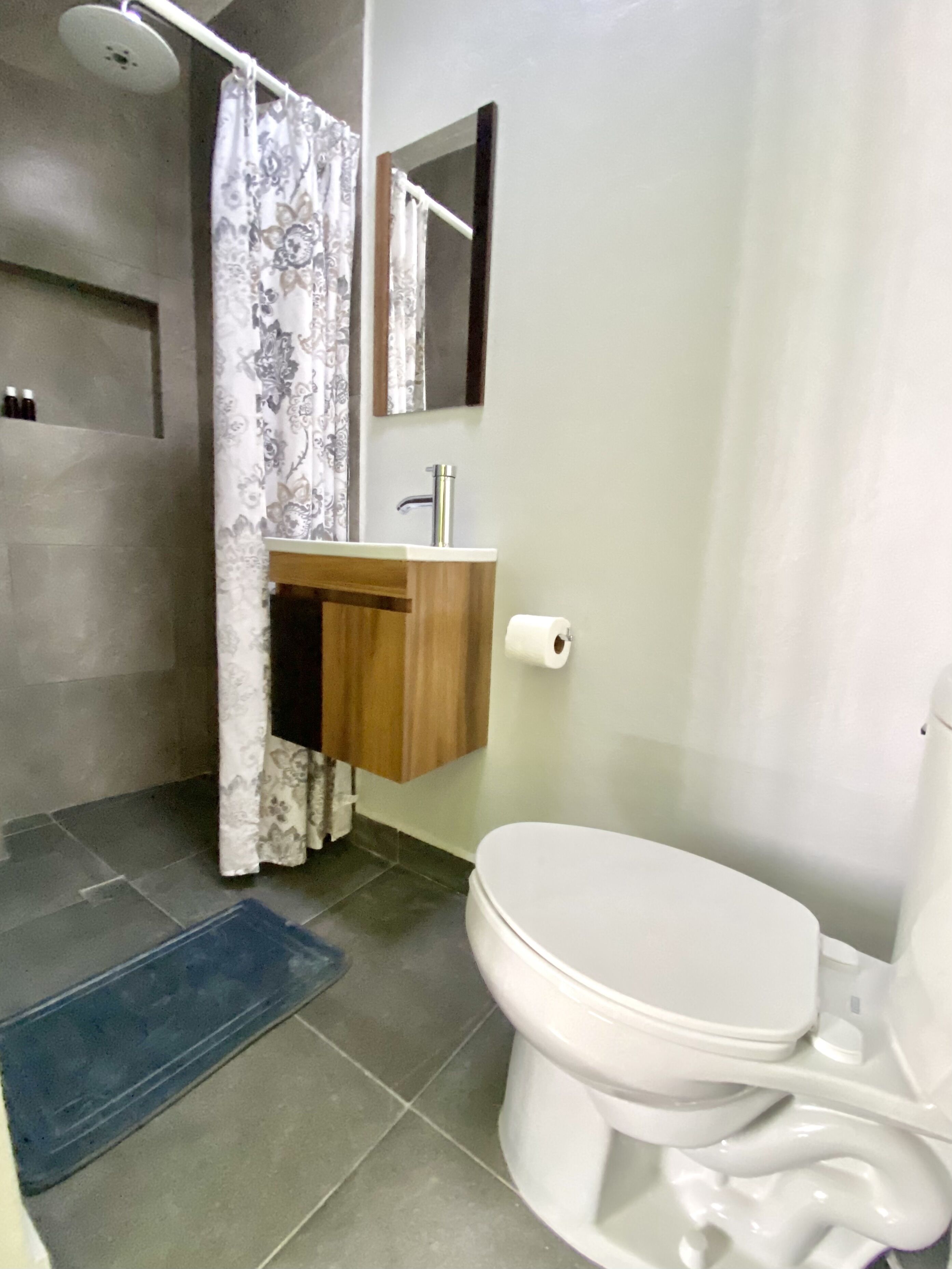 Une salle de bain avec des toilettes, un lavabo avec un miroir au-dessus, un rideau de douche et un tapis bleu sur le sol.