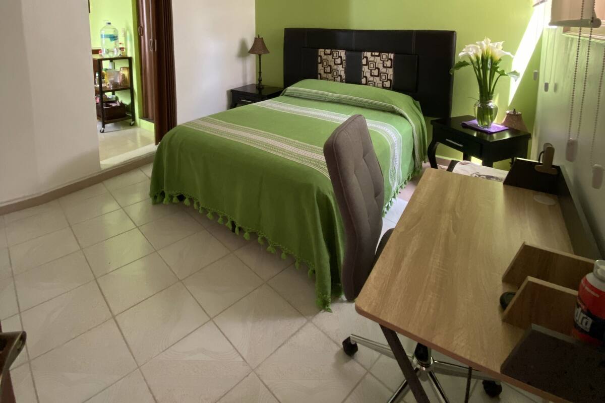 Un dormitorio con cama, escritorio, silla, ventilador de techo y mesita de noche con lámpara.