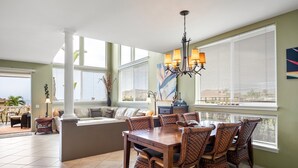 A dining area with a wooden table and wicker chairs, large windows with blinds, and a chandelier. - Alii Cove I-4 in Historic Kailua Village (Kailua Kona)
