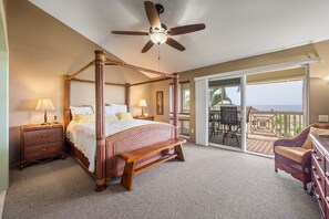 Ein Bett mit Baldachin, ein Deckenventilator, ein Nachttisch mit Lampe, ein Sessel und Blick auf den Ozean durch eine Glastür.