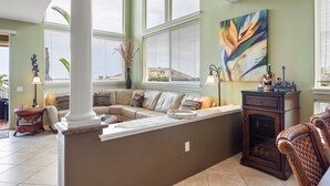 A spacious living room with a beige sofa, a coffee table, and a large window with blinds. - Alii Cove I-4 in Historic Kailua Village (Kailua Kona)