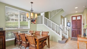 A dining area with a wooden table and wicker chairs, a staircase with white railings, and a door with a decorative vase. - Alii Cove I-4 in Historic Kailua Village (Kailua Kona)