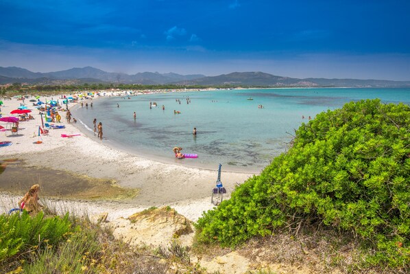 A beach with clear turquoise water, people swimming and sunbathing, and a view of distant mountains.