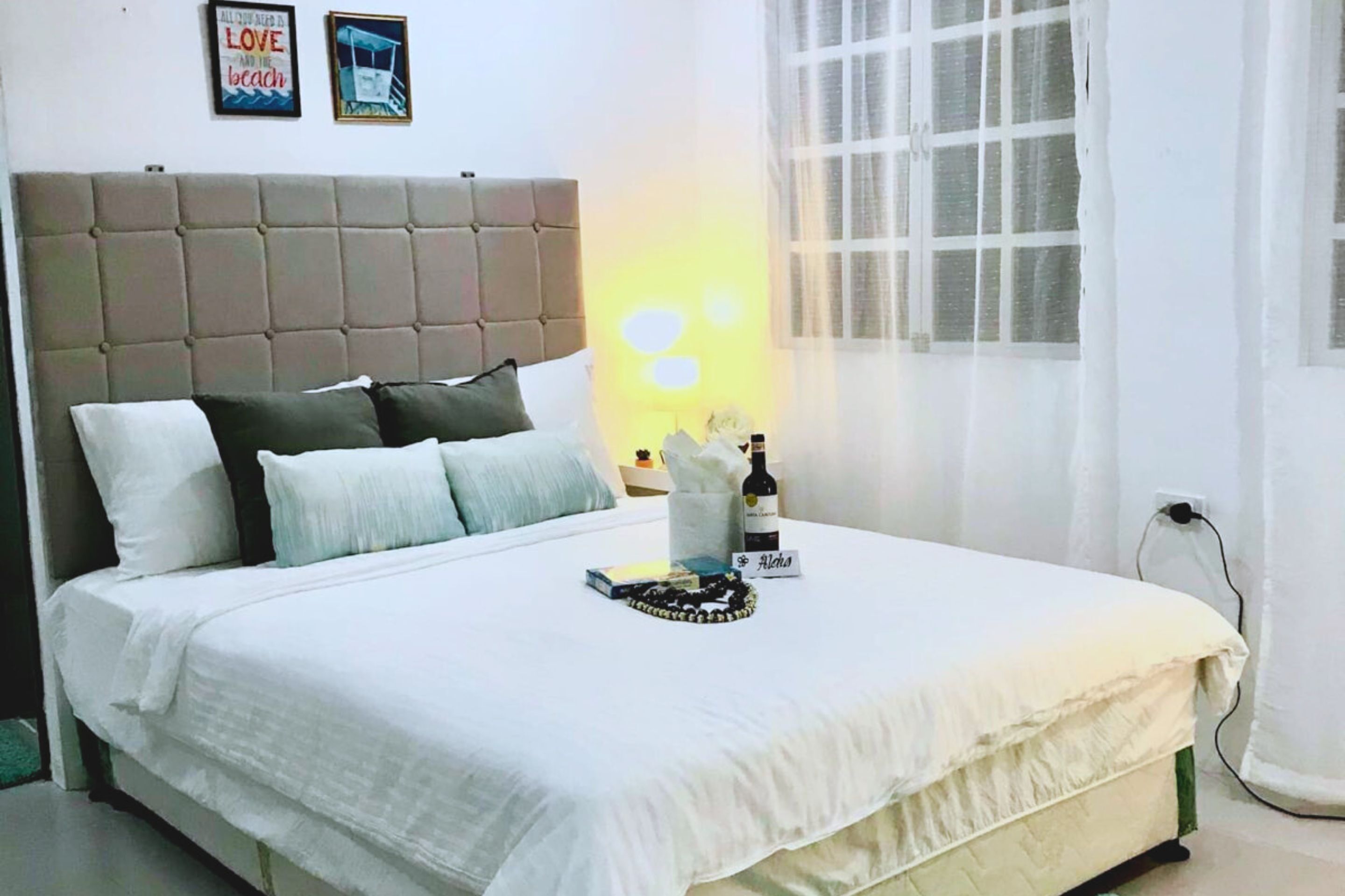 A neatly made bed with a white comforter and pillows, a bedside table with a bottle and a book, and a wall with two framed pictures.
