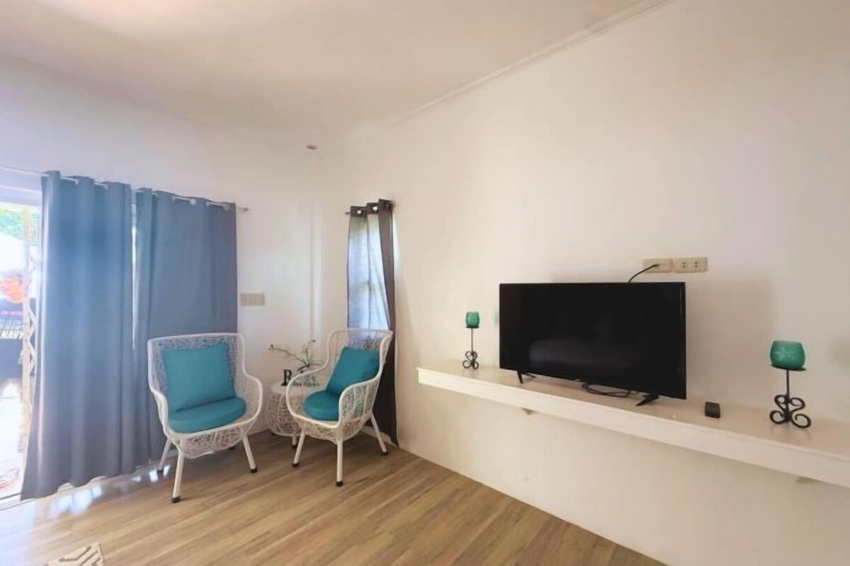 Una sala de estar con un televisor de pantalla plana sobre un estante blanco, dos sillones de mimbre con cojines azul turquesa y una mesita con un adorno.