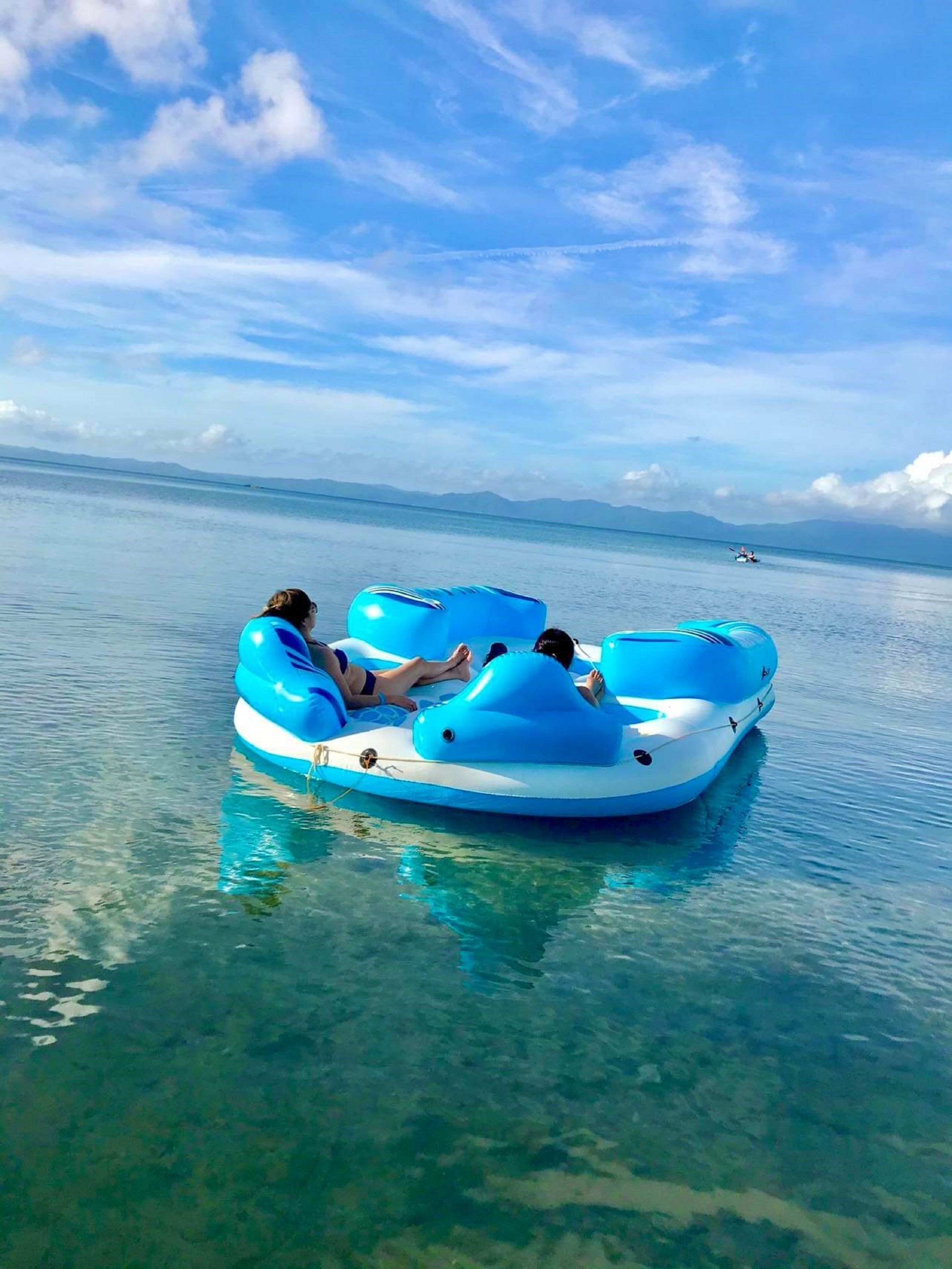 A clear blue sky with a few clouds, a calm sea, and a person in a blue inflatable raft.