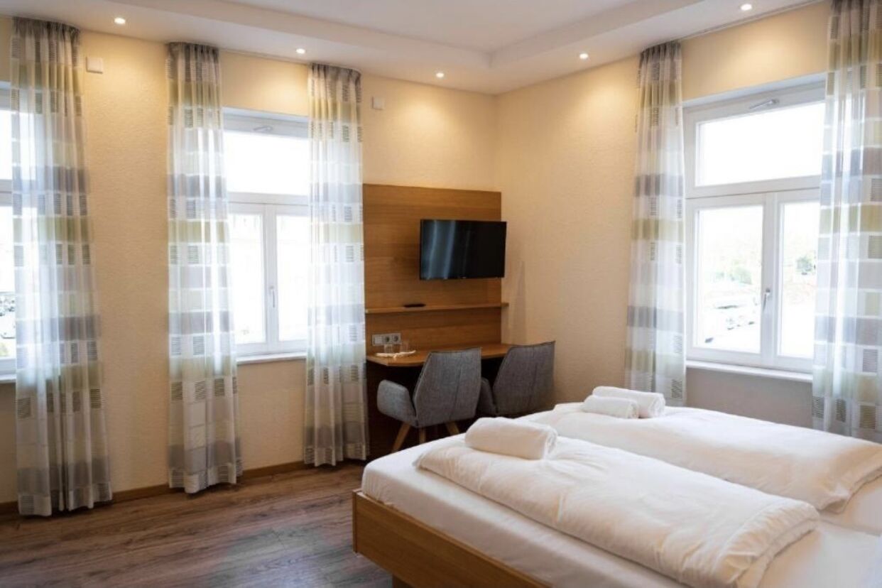 A hotel room with a bed, a desk with a chair, a television, and large windows with curtains.