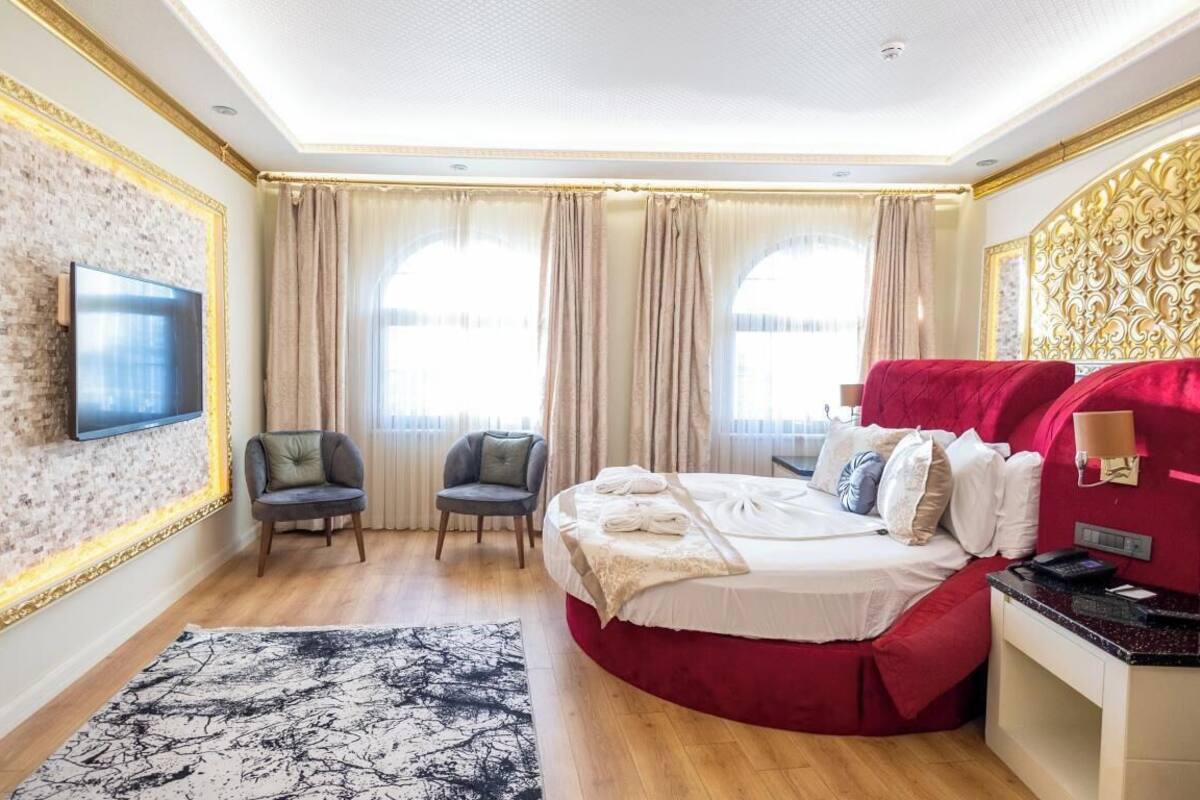 Habitació d'hotel amb un llit gran, una televisió penjada a una paret amb relleu, dues cadires, un escriptori amb un ordinador i un gran finestral amb cortines.