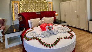 Quarto de hotel com cama redonda, cabeceira vermelha e painel decorativo na parede. Há uma pequena mesa com luminária e uma cadeira. O quarto tem piso de madeira e armários brancos.