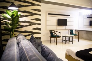 A modern hotel room with a sofa, a dining table, two chairs, a potted plant, and a flat-screen TV mounted on the wall.