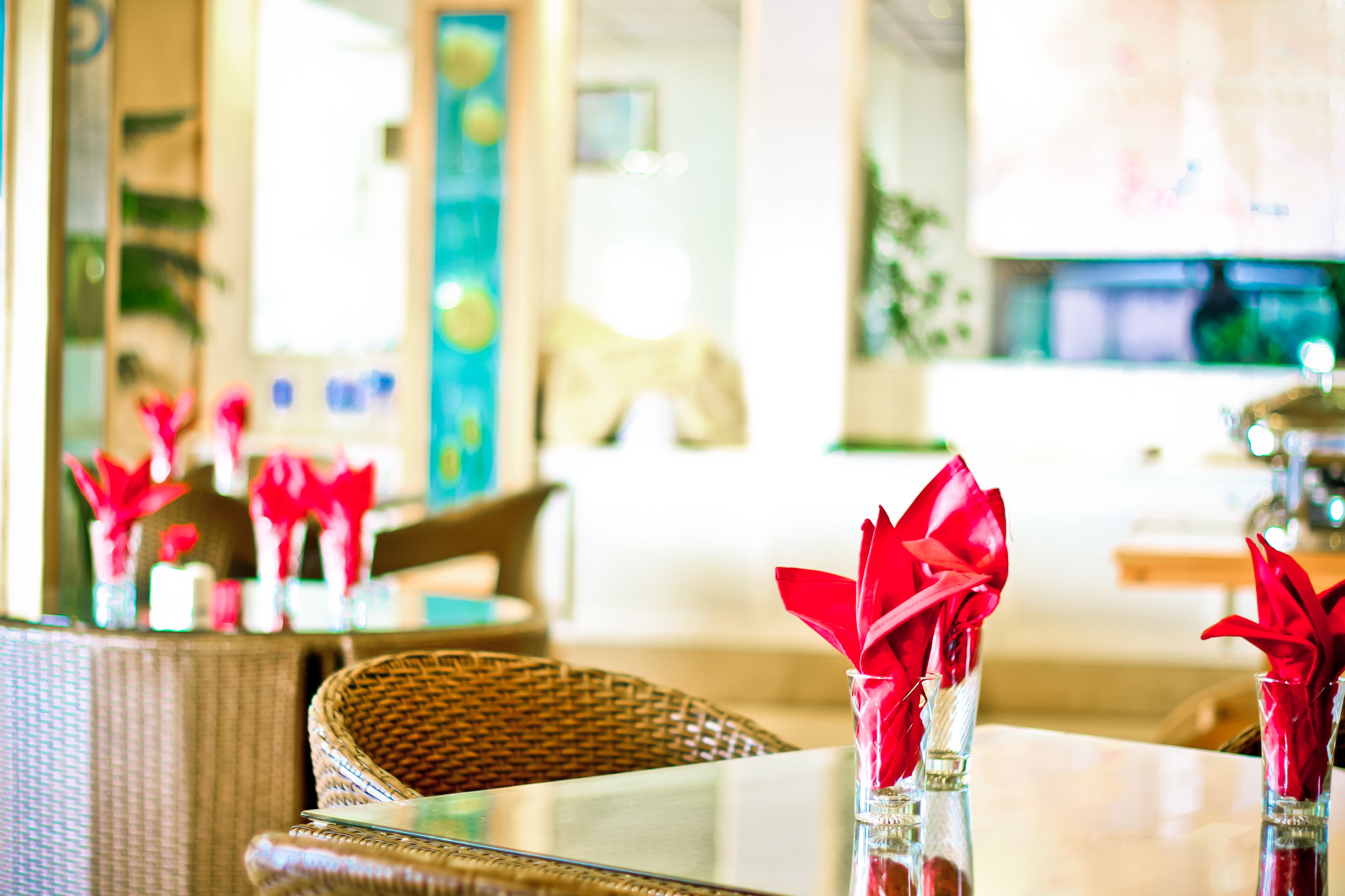 A dining area with wicker furniture and glass tables, featuring red napkins in clear vases.