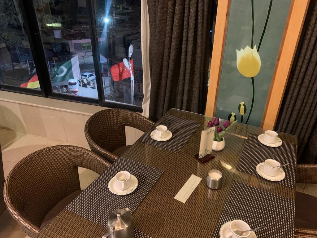 Een eethoek met een tafel gedekt voor vier personen, inclusief witte borden, bekers en bestek. Er staan rieten stoelen rond de tafel, en een raam met uitzicht op een straatbeeld.