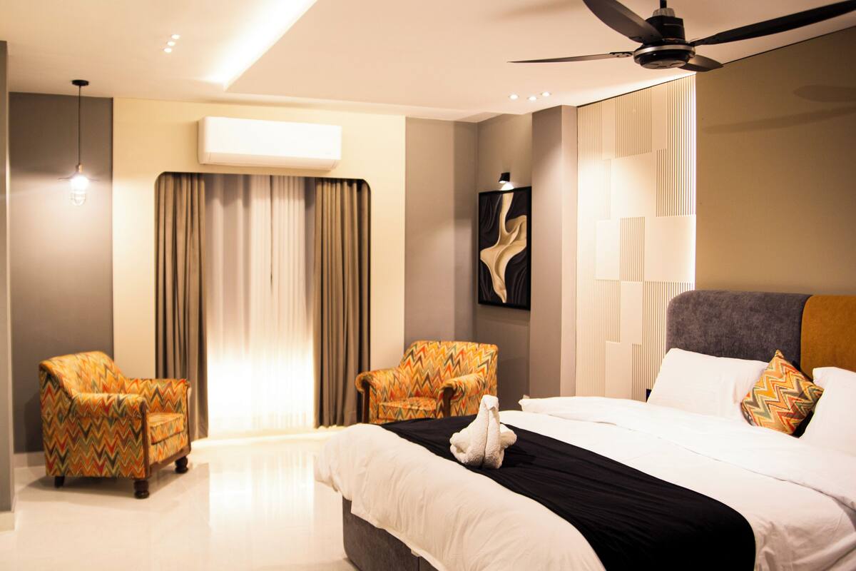 A hotel room with a bed, two armchairs, a ceiling fan, an air conditioning unit, and a painting on the wall.
