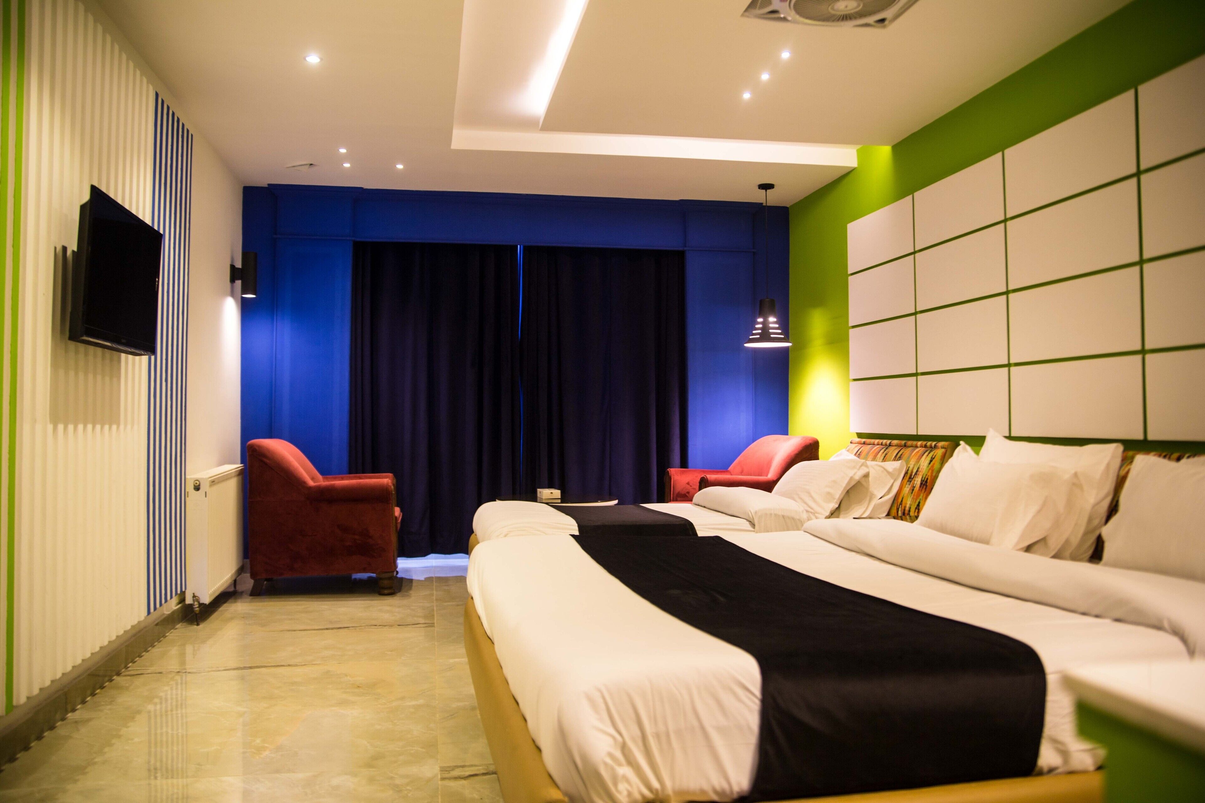 A hotel room with a large bed, two red armchairs, a flat-screen TV mounted on the wall, and a large window with curtains.