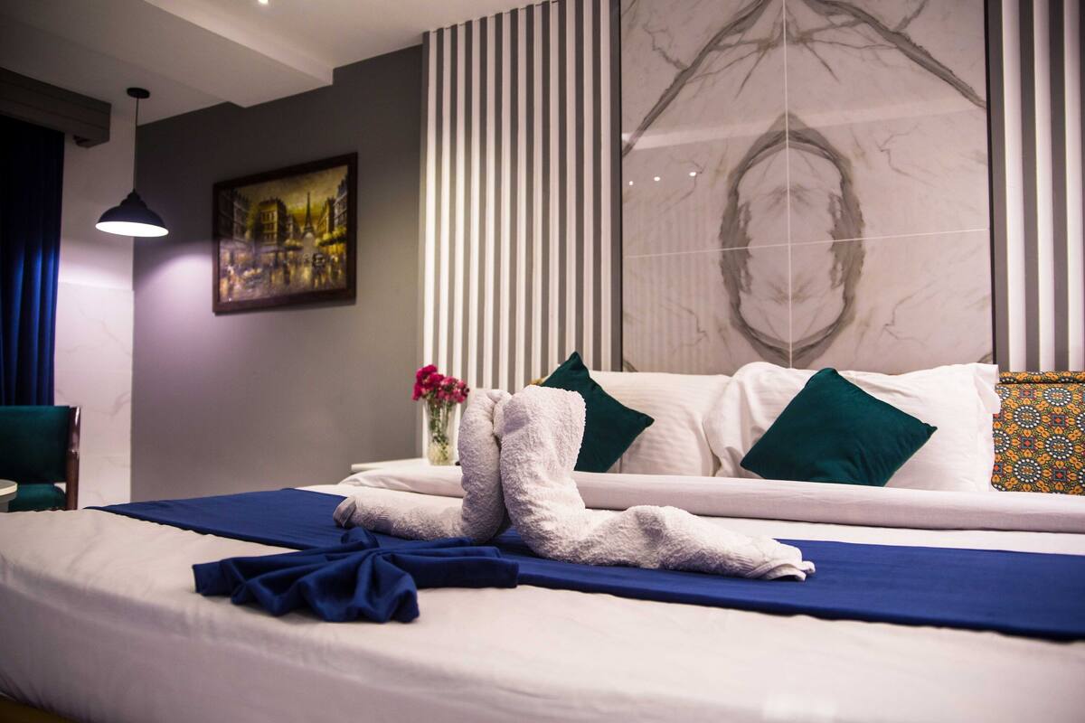 A neatly made bed with a blue bedspread and white towels, a green chair, a painting on the wall, and a large mirror.