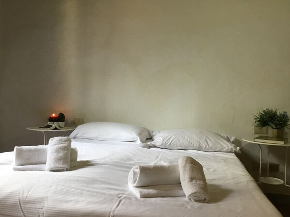A neatly made bed with white linens and rolled towels, a bedside table with a small plant, and a nightstand with a candle and a book.