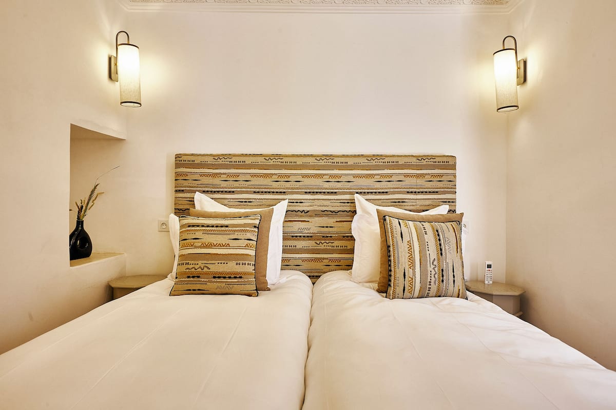 A double bed with patterned pillows and headboard, two bedside tables with lamps, and a vase of dried plants on a nightstand.