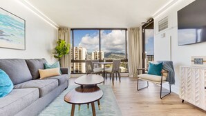 Un moderno salón con un sofá gris, una pequeña mesa redonda y una zona de comedor con sillas. Hay un ventanal con vistas a edificios y montañas.