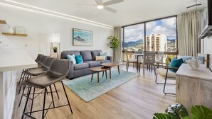 Una sala de estar moderna con un sofá gris, un comedor con taburetes de bar, un ventanal con vista a la ciudad y un ventilador de techo.