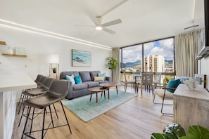 A modern living room with a grey sofa, a dining area with bar stools, a large window with a city view, and a ceiling fan. - Newly renovated, Free Parking, One block to Waikiki Beach (Honolulu)