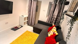 A modern living room with a grey sofa, a yellow rug, and a TV mounted on the wall. - Delightful holiday home away from home, wetroom, UFH, garden/parking, local park (London)