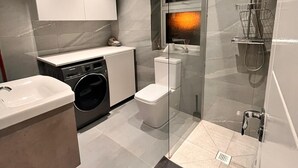 A modern bathroom with a glass shower enclosure, a built-in washing machine, and a toilet. - Delightful holiday home away from home, wetroom, UFH, garden/parking, local park (London)