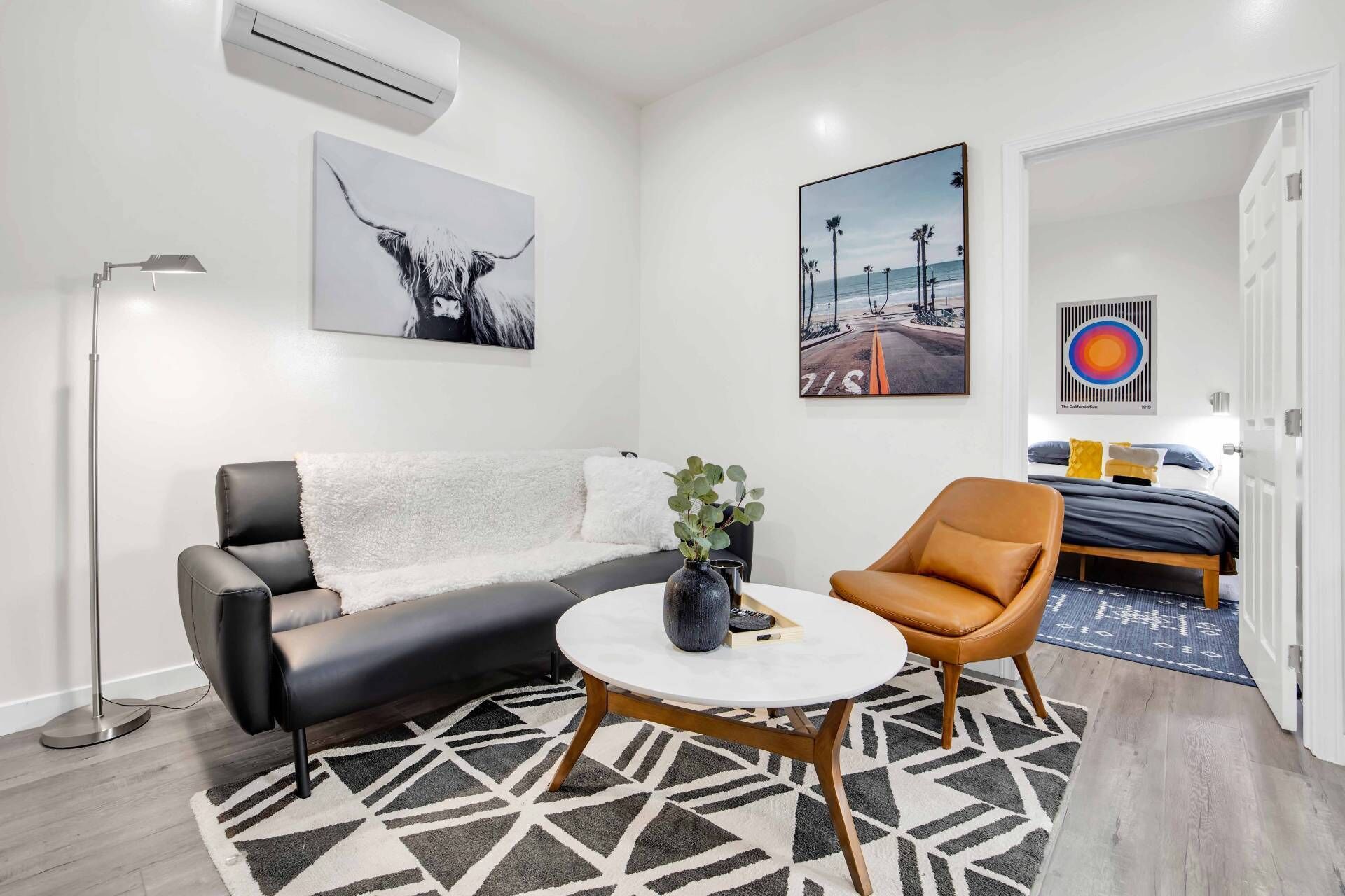 Een moderne woonkamer met een zwarte leren bank, een ronde salontafel en een bruine leren fauteuil. Er hangen drie kunstwerken aan de muren, een witte airconditioner en een deur naar een andere kamer.