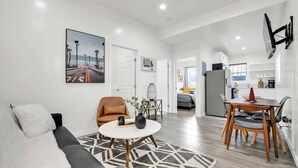 Un salon moderne avec un canapé, une table basse et un coin repas comprenant une table et des chaises. Au fond, on aperçoit une cuisine avec un réfrigérateur et une fenêtre.