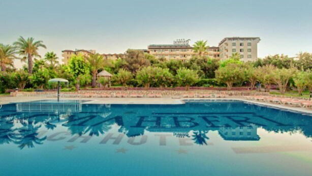 Una piscina con la parola "ZAHER" riflessa sull'acqua, circondata da vegetazione e un edificio sullo sfondo.