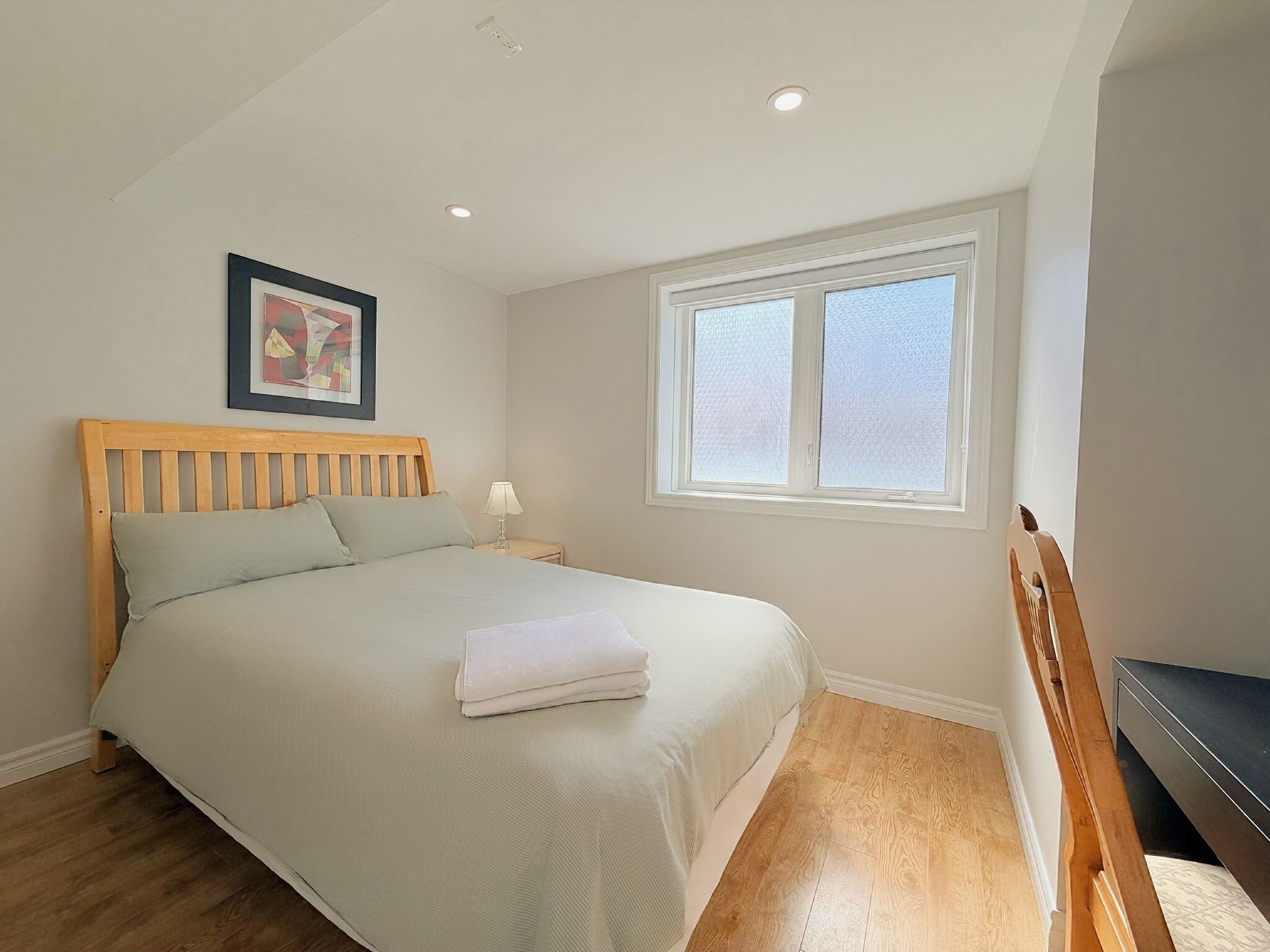 Un dormitorio con una cama de madera, una mesita de noche, una lámpara, una ventana y un cuadro enmarcado en la pared.