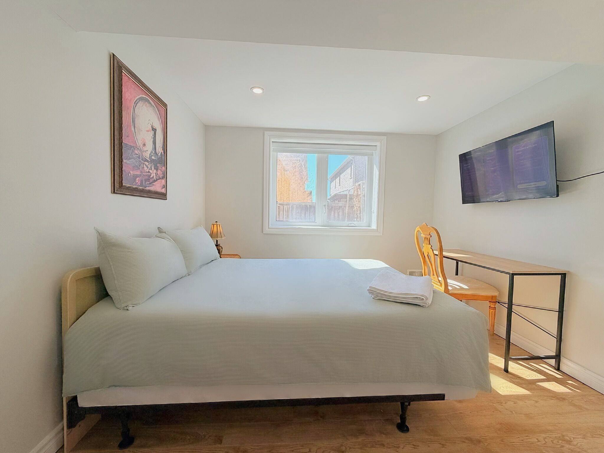 A bedroom with a bed, a desk, a television, a window, and a painting.