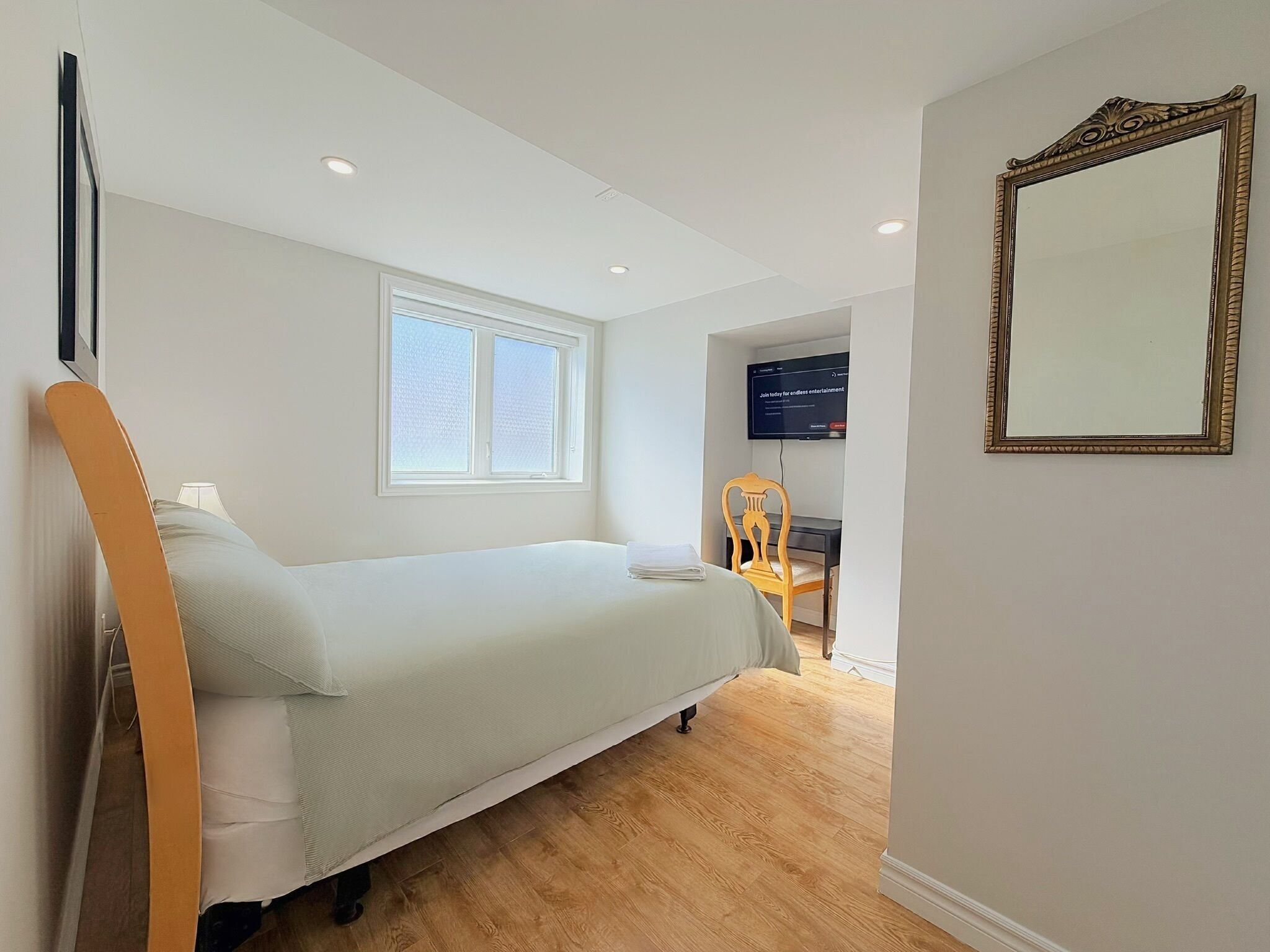 Un dormitorio con cama, ventana, escritorio, silla, televisor y espejo.